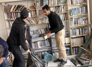Ketika Gaza Baik-baik Saja: Buku Selamatkan Memori Gaza Utuh 1 Dua pemuda sedang mengevakuasi buku-buku dari rak ke dalam kantong plastik hitam besar di dalam ruangan perpustakaan yang hancur dipenuhi puing-puing bangunan dan atap yang runtuh.