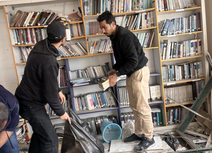 Dua pemuda sedang mengevakuasi buku-buku dari rak ke dalam kantong plastik hitam besar di dalam ruangan perpustakaan yang hancur dipenuhi puing-puing bangunan dan atap yang runtuh.