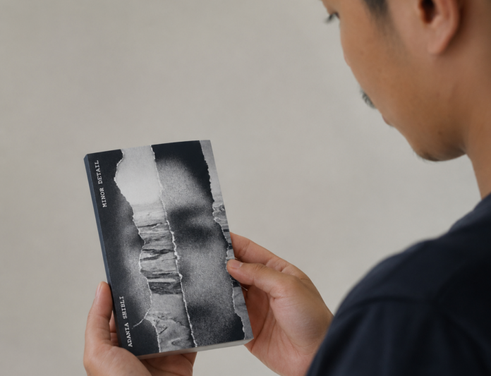 Seorang pria terlihat dari sisi belakang sedang membaca buku kecil bercover hitam putih bertuliskan Minor Detail karya Adania Shibli. Wajah pria hanya tampak sebagian dalam posisi profil.
